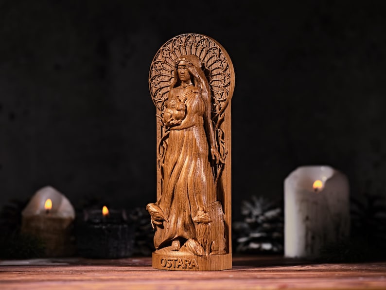 May include: A wooden statue of a woman holding a bundle, with the word "OSTARA" at the base. The statue is intricately carved with a detailed design, and is set against a dark background with lit candles.