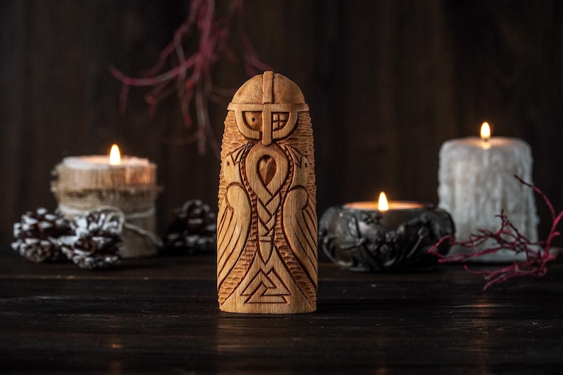 May include: A wooden carving of a Viking warrior with a helmet, beard, and two birds on either side. The carving is on a dark wooden surface with three lit candles in the background.