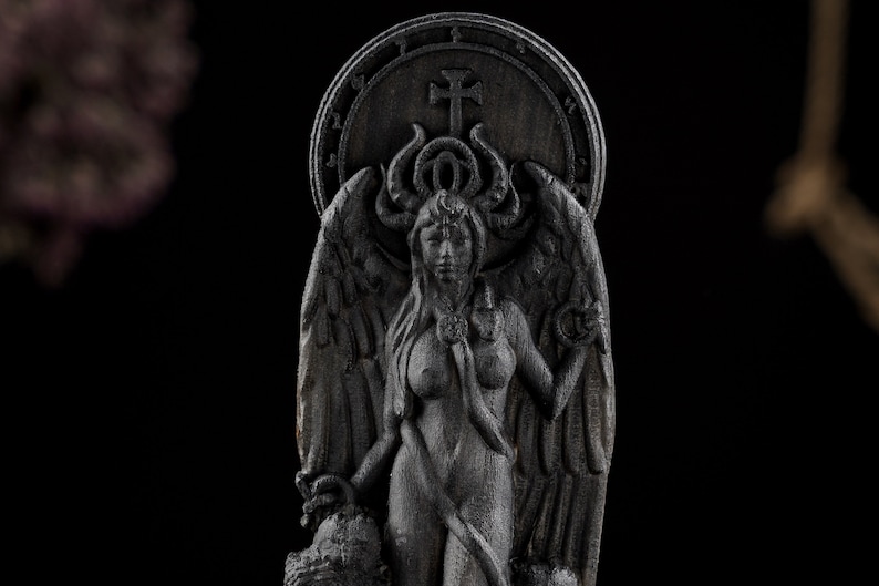 May include: A dark gray sculpture of a female figure with wings, horns, and a halo. The figure is nude and holds a staff. The sculpture has a cross on the halo and is set against a dark background.