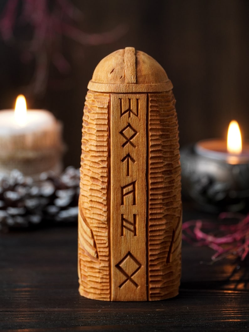 May include: A wooden box with a carved lid and runes on the front. The box is sitting on a dark wooden surface with two candles in the background.