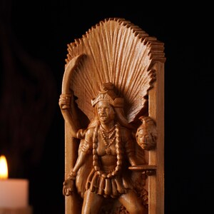 Pu&ograve; includere: Un intaglio in legno marrone chiaro raffigurante una figura dalle braccia multiple con un copricapo prominente e un'aureola a ventaglio. La figura tiene una lama curva in una mano e una testa mozzata nell'altra, su sfondo scuro.