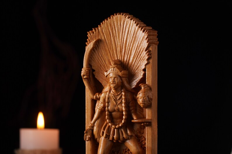 Pu&ograve; includere: Un intaglio in legno marrone chiaro raffigurante una figura dalle braccia multiple con un copricapo prominente e un'aureola a ventaglio. La figura tiene una lama curva in una mano e una testa mozzata nell'altra, su sfondo scuro.
