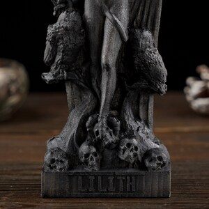May include: A dark gray statue of Lilith, featuring intricate details. The statue depicts Lilith with owls and skulls, with the name "LILITH" at the base. The statue is on a wooden surface.