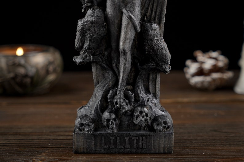 May include: A dark gray statue of Lilith, featuring intricate details. The statue depicts Lilith with owls and skulls, with the name "LILITH" at the base. The statue is on a wooden surface.
