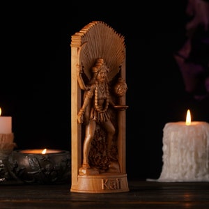 Pu&ograve; includere: Una statua di legno della dea ind&ugrave; Kali, in piedi in una cornice intagliata. La statua &egrave; dipinta di un colore marrone scuro e presenta dettagli intricati. La dea tiene un teschio in una mano e una spada nell'altra. La statua &egrave; posizionata su uno sfondo scuro con due candele su entrambi i lati.