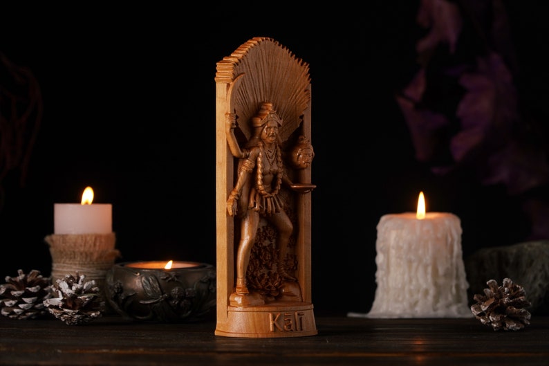 Pu&ograve; includere: Una statua di legno della dea ind&ugrave; Kali, in piedi in una cornice intagliata. La statua &egrave; dipinta di un colore marrone scuro e presenta dettagli intricati. La dea tiene un teschio in una mano e una spada nell'altra. La statua &egrave; posizionata su uno sfondo scuro con due candele su entrambi i lati.
