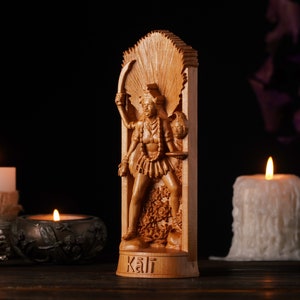 Pu&ograve; includere: Una statua in legno della dea ind&ugrave; Kali, intagliata con dettagli. &Egrave; raffigurata con pi&ugrave; braccia, che tengono una spada e una testa mozzata. La statua &egrave; ambientata su uno sfondo scuro con candele accese e pigne.