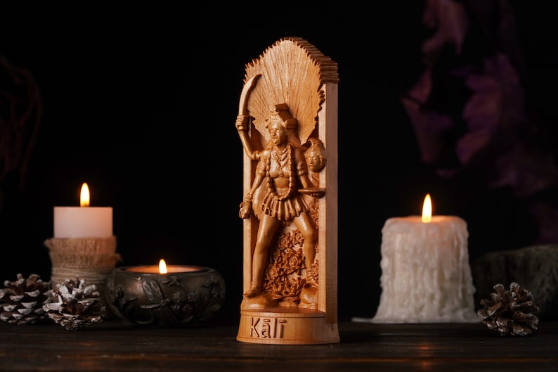 Pu&ograve; includere: Una statua in legno della dea ind&ugrave; Kali, intagliata con dettagli. &Egrave; raffigurata con pi&ugrave; braccia, che tengono una spada e una testa mozzata. La statua &egrave; ambientata su uno sfondo scuro con candele accese e pigne.