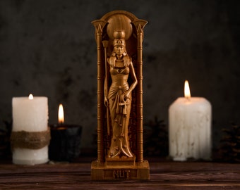 Puede incluir: Una estatua de madera de la diosa egipcia Nut, de pie en un marco de madera tallado. La estatua está pintada de oro y presenta detalles intrincados. La estatua está rodeada de velas y piñas.
