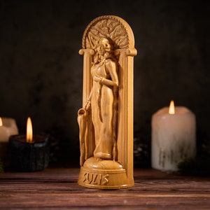 May include: A wooden statue of a woman in a flowing robe, standing in a carved archway. The statue is on a wooden base with the word "Sulis" inscribed on it. The statue is surrounded by candles.