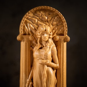 May include: A wooden sculpture of a woman in a flowing robe, standing in a doorway with a decorative archway. The sculpture is carved in a light brown wood and has a detailed design.