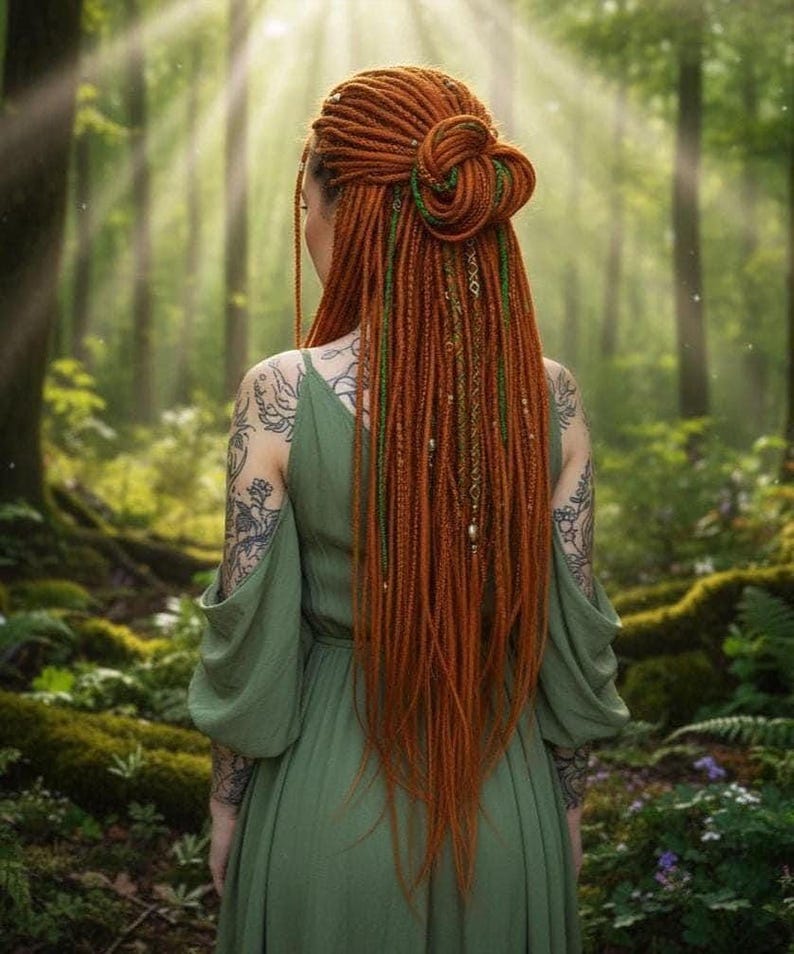 A woman with long, vibrant red braids and tattoos stands in a lush, green forest, her back to the camera.