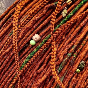 a close-up view of a collection of braided hair strands in various shades of orange and green, with small green beads and a gold-colored charm attached to some of the braids.