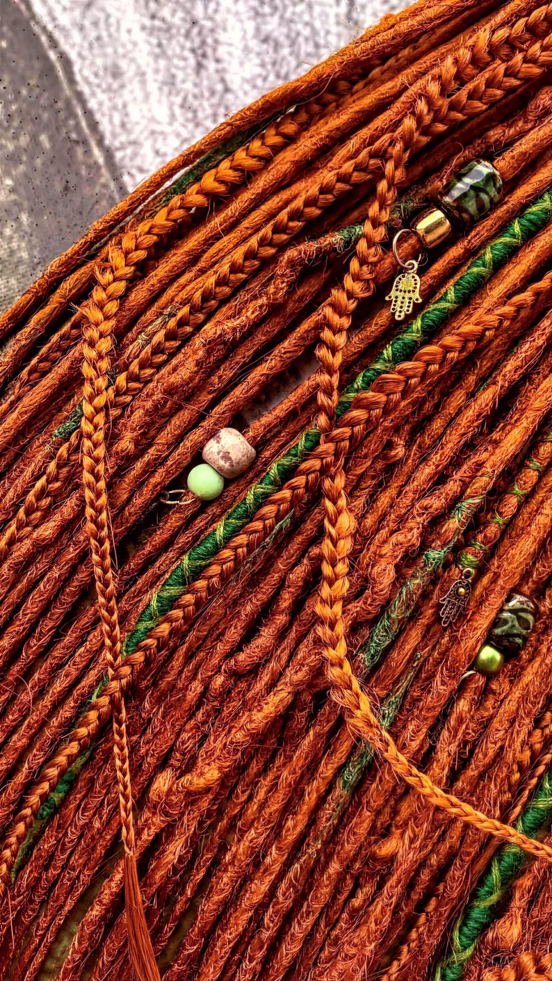a close-up view of a collection of braided hair strands in various shades of orange and green, with small green beads and a gold-colored charm attached to some of the braids.