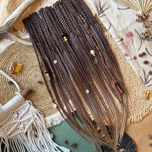 Op de afbeelding: Een bundel bruine en blonde synthetische dreadlocks, versierd met kralen en decoratieve accenten. De dreadlocks liggen op een getextureerd oppervlak met een macramé tas en decoratieve elementen.