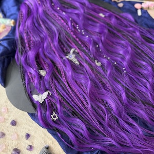 a close-up view of a person's long, vibrant purple hair adorned with various accessories, including a Star of David charm and small silver charms. The hair is styled in a braided manner, giving it a unique and eye-catching appearance. The background is a dark blue fabric, which contrasts with the bright purple hair and adds depth to the image.
