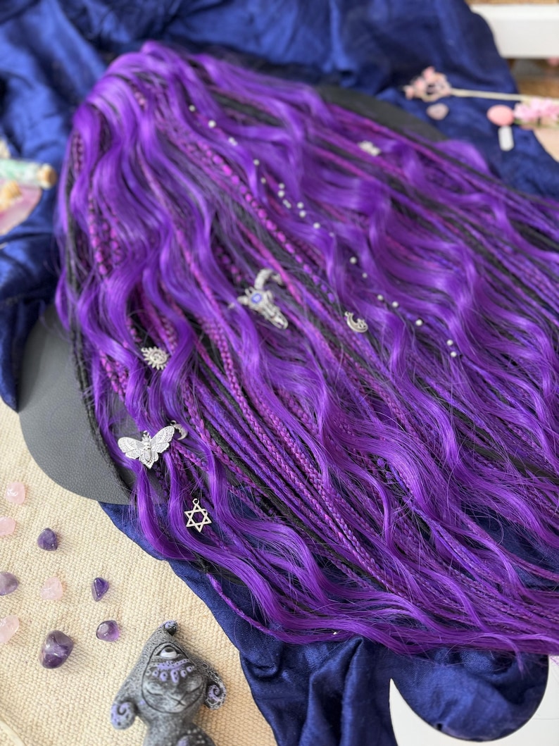 a close-up view of a person's long, vibrant purple hair adorned with various accessories, including a Star of David charm and small silver charms. The hair is styled in a braided manner, giving it a unique and eye-catching appearance. The background is a dark blue fabric, which contrasts with the bright purple hair and adds depth to the image.