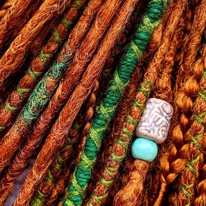a close-up view of a collection of braided hair strands, with some of the strands being wrapped in green and orange yarn. There is a small blue bead attached to one of the braids, adding a decorative element to the arrangement.