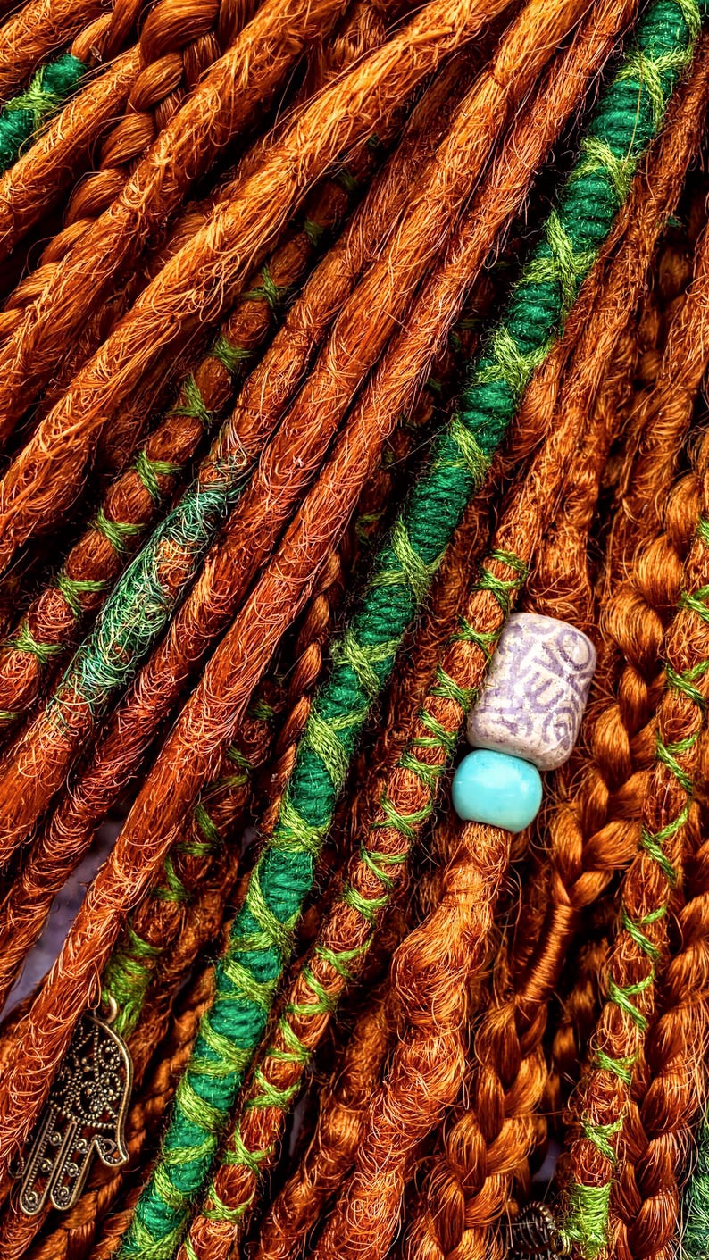 a close-up view of a collection of braided hair strands, with some of the strands being wrapped in green and orange yarn. There is a small blue bead attached to one of the braids, adding a decorative element to the arrangement.