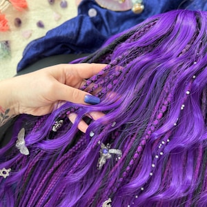 A hand is holding a section of long, purple braided hair with various accessories, such as silver and purple charms, and a blue fabric in the background.