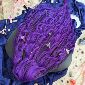 a large, vibrant purple wig with intricate braids and various accessories, including a black object, scattered around it on a blue fabric surface.