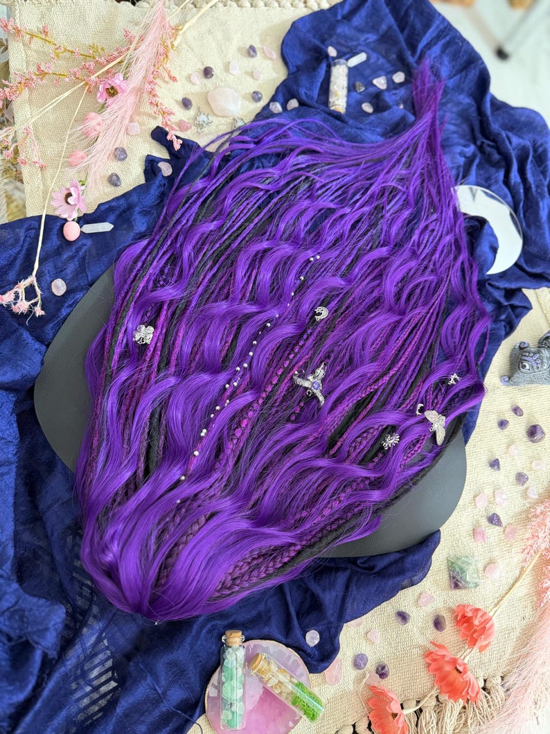 a large, vibrant purple wig with intricate braids and various accessories, including a black object, scattered around it on a blue fabric surface.
