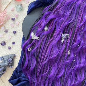 a close-up view of a person's long, vibrant purple hair adorned with various accessories, including silver and gemstone embellishments. The hair is styled in a way that highlights its length and color, creating a striking visual effect. The background is a mix of natural and artificial elements, including flowers, gemstones, and a blue fabric, adding depth and texture to the overall composition.