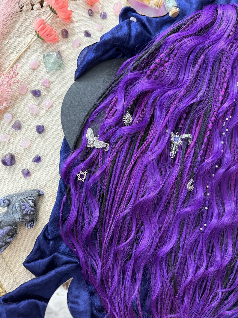 a close-up view of a person's long, vibrant purple hair adorned with various accessories, including silver and gemstone embellishments. The hair is styled in a way that highlights its length and color, creating a striking visual effect. The background is a mix of natural and artificial elements, including flowers, gemstones, and a blue fabric, adding depth and texture to the overall composition.