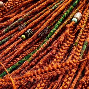 a close-up view of a collection of braided hair strands in various shades of orange and green, with some beads or accessories attached to the braids.