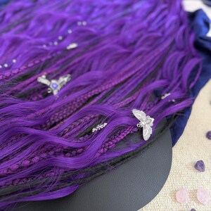 a close-up view of a vibrant purple wig adorned with silver butterfly-shaped earrings, resting on a surface with scattered gemstones.