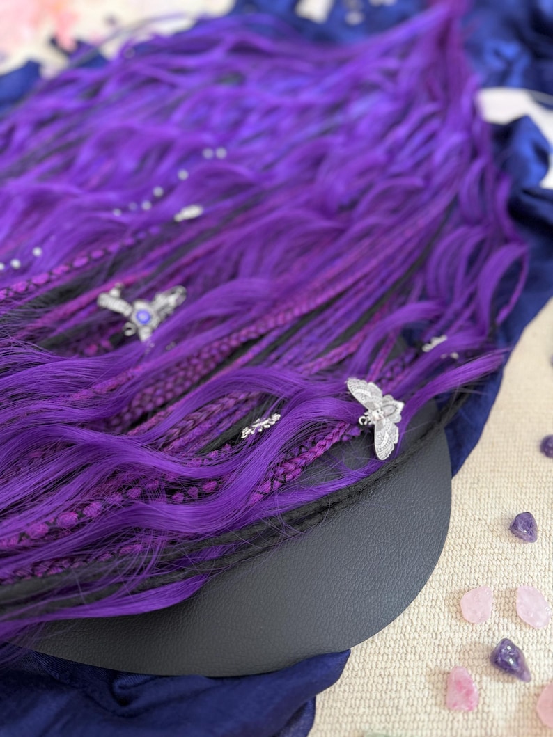 a close-up view of a vibrant purple wig adorned with silver butterfly-shaped earrings, resting on a surface with scattered gemstones.
