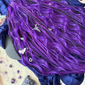 a close-up view of a person's long, vibrant purple hair adorned with various accessories, including small silver charms and gemstones. The hair appears to be styled in a braided manner, with the ends of the hair falling over the edge of the frame. The background is a dark blue fabric, providing a striking contrast to the bright purple hair.