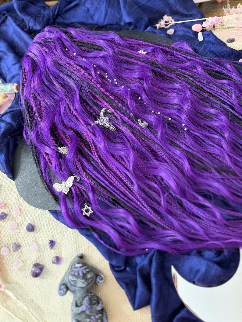 a close-up view of a person's long, vibrant purple hair adorned with various accessories, including small silver charms and gemstones. The hair appears to be styled in a braided manner, with the ends of the hair falling over the edge of the frame. The background is a dark blue fabric, providing a striking contrast to the bright purple hair.