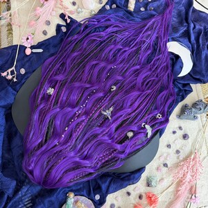 a large, vibrant purple wig with intricate braids and various accessories scattered around it, including crystals, flowers, and other decorative elements.