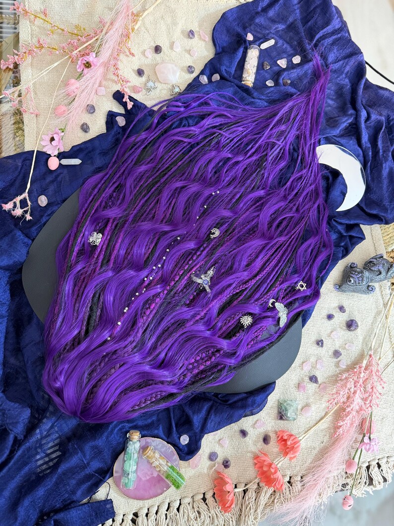 a large, vibrant purple wig with intricate braids and various accessories scattered around it, including crystals, flowers, and other decorative elements.