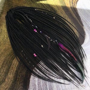Puede incluir: Un conjunto de extensiones de cabello negro con numerosos mechones finos trenzados. Las extensiones están adornadas con pequeñas cuentas metálicas, anillos plateados y una pluma de color púrpura y rosa vibrante en un mechón.