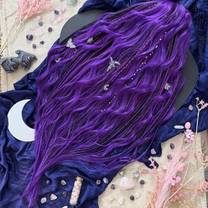a purple wig with long, wavy hair, surrounded by various objects such as crystals, flowers, and a crescent moon.