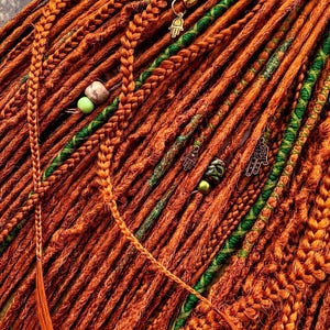 a close-up view of a collection of braided hair strands in various shades of orange and green, with small green beads and charms attached to the braids.
