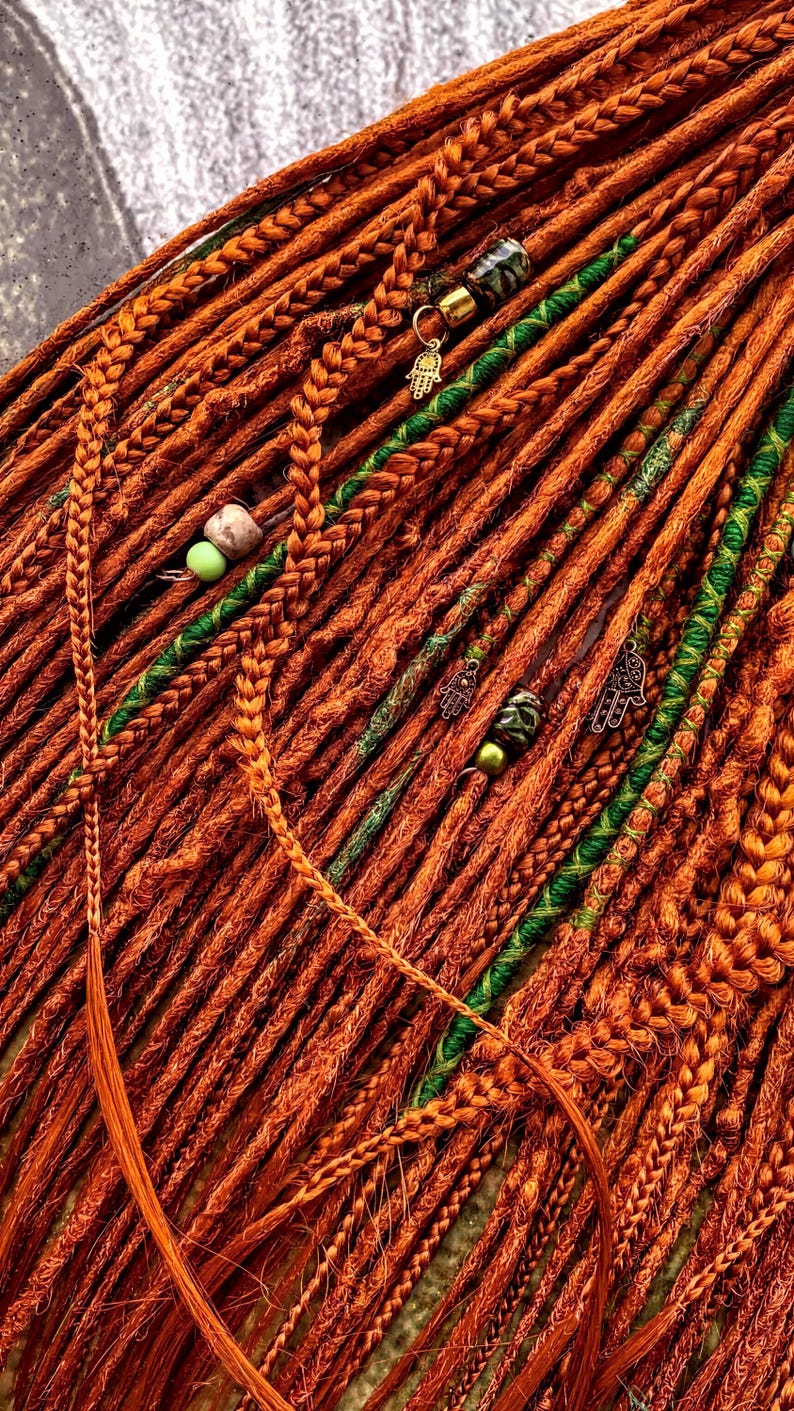 a close-up view of a collection of braided hair strands in various shades of orange and green, with small green beads and charms attached to the braids.