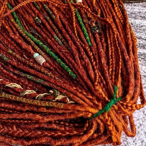 a close-up view of a large pile of braided hair strands, with some of the strands adorned with green and white beads. The hair appears to be of a reddish-brown color, and the beads are scattered throughout the strands, adding a decorative element to the overall appearance.