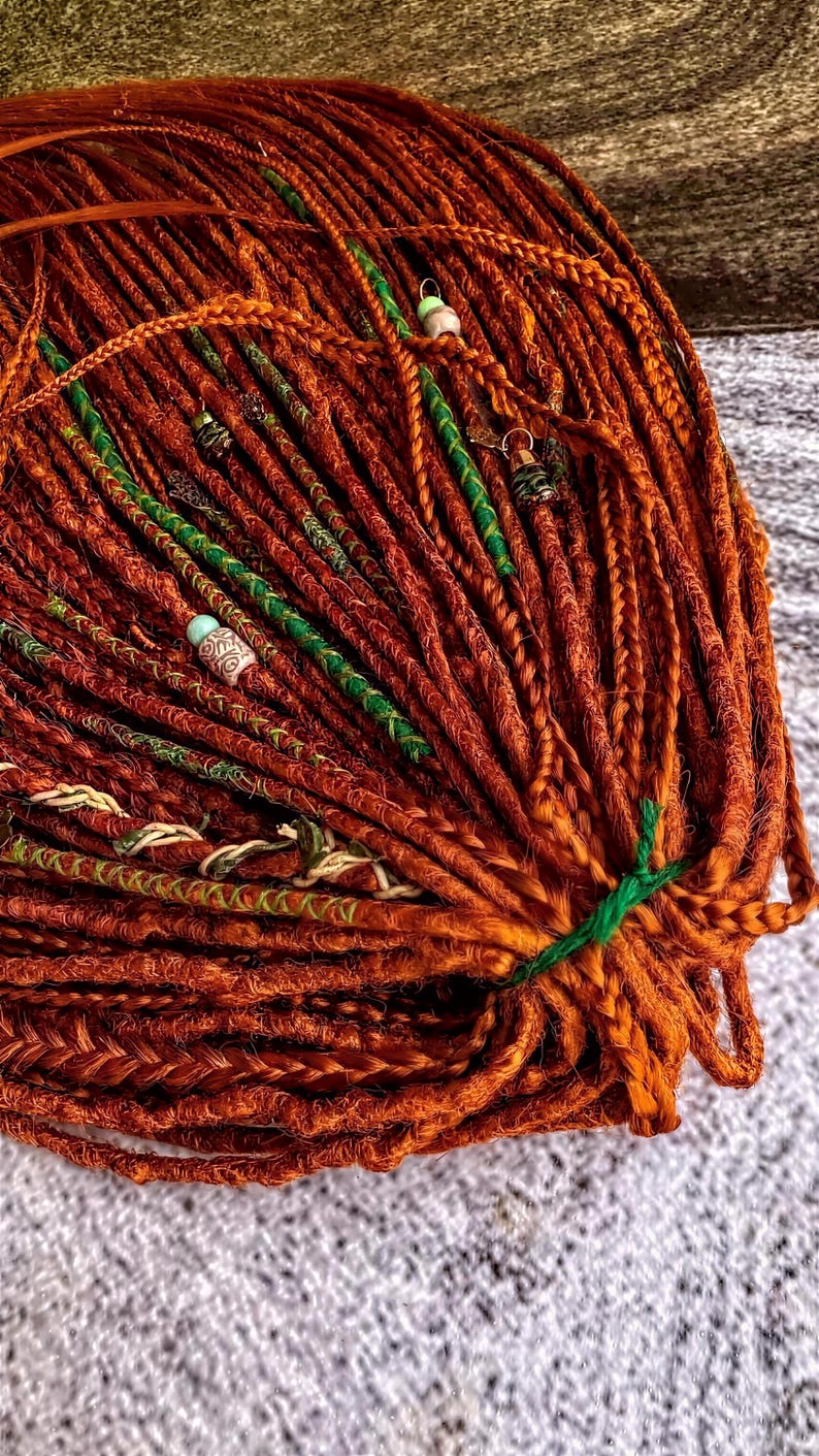 a close-up view of a large pile of braided hair strands, with some of the strands adorned with green and white beads. The hair appears to be of a reddish-brown color, and the beads are scattered throughout the strands, adding a decorative element to the overall appearance.