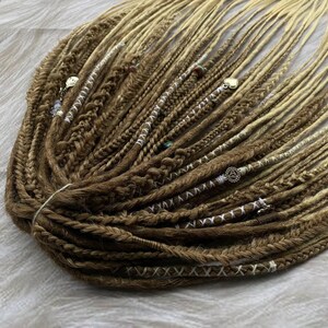 May include: A bundle of brown and blonde synthetic dreadlocks. The dreads are intricately braided and decorated with beads and charms. The color gradient transitions from a dark brown at the roots to a light blonde at the tips. The dreads are styled and ready to wear.