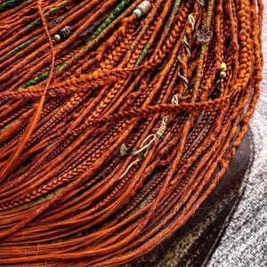a close-up view of a large pile of braided orange hair, with various green and silver beads and accessories attached to the braids.