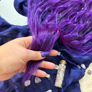 A hand holding a purple hair accessory, surrounded by crystals and a crescent moon on a blue fabric background.