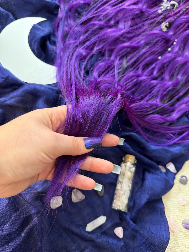 A hand holding a purple hair accessory, surrounded by crystals and a crescent moon on a blue fabric background.