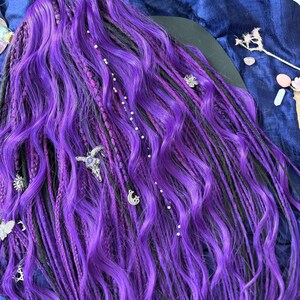 a close-up view of a person's long, vibrant purple braided hairstyle. The hair is styled in intricate braids and adorned with various decorative elements, such as small charms and beads. The overall color and style of the hair suggest it is a unique and eye-catching hairstyle.