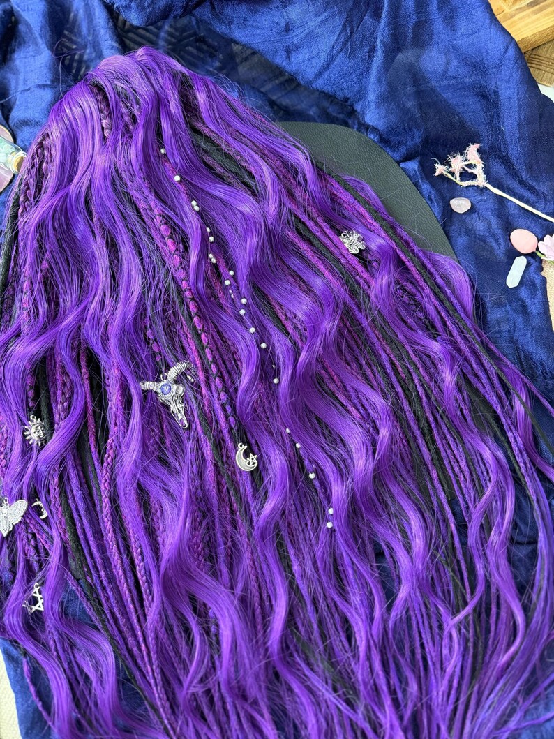 a close-up view of a person's long, vibrant purple braided hairstyle. The hair is styled in intricate braids and adorned with various decorative elements, such as small charms and beads. The overall color and style of the hair suggest it is a unique and eye-catching hairstyle.