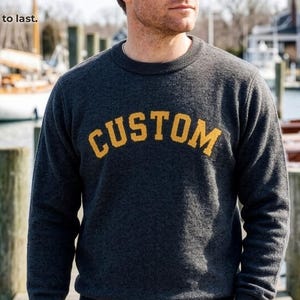 May include: A dark gray long-sleeve knit sweater with the word CUSTOM in yellow block letters across the chest. The text A real knit sweater. Not a sweatshirt. Custom text, made to last. is visible in the upper left corner of the image.