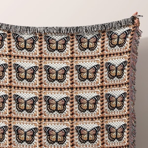 May include: A woven throw blanket featuring a repeating pattern of monarch butterflies in shades of orange, black, and white. The blanket has a fringed edge and is held up against a neutral background. The butterflies are arranged in a grid-like pattern.