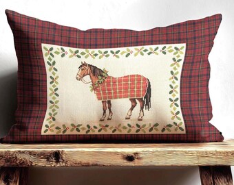 May include: Rectangular cushion with a red tartan pattern. The centre displays a horse wearing a red plaid blanket and a wreath. The horse is set against a cream background with a border of green holly leaves. The cushion is on a wooden bench.
